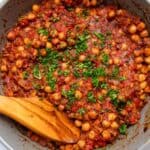 Red stewed chickpeas topped with parsley in a gray pot with a wooden spatula resting on the chickpeas.