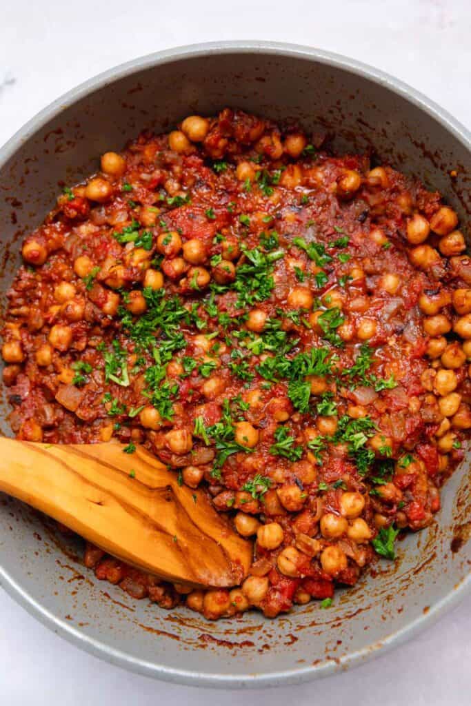 Red stewed chickpeas topped with parsley in a gray pot with a wooden spatula resting on the chickpeas.