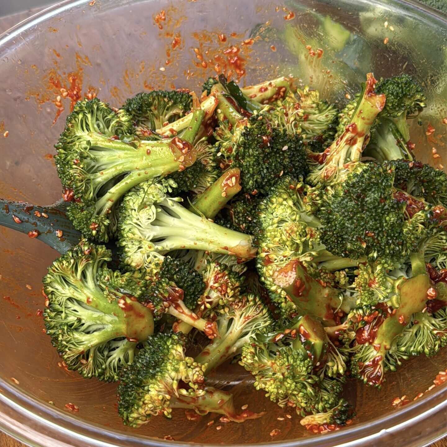 Broccoli florets tossed in a gochujang and oil in a large glass bowl on a wooden countertop.