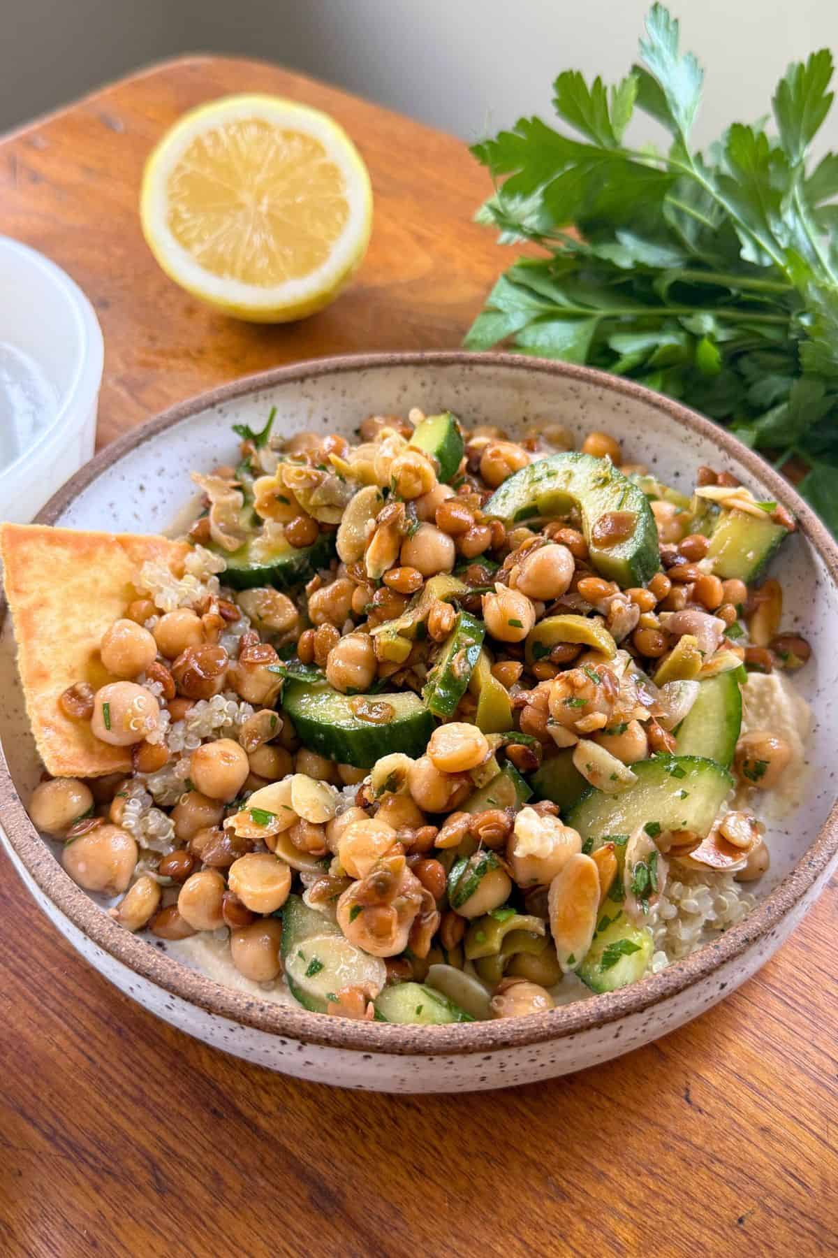 Brown-rimmed white bowl filled with lentils and chickpeas on a wood table with parsley and half of a lemon in the background.