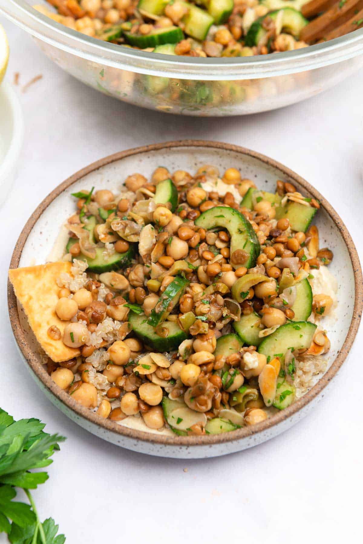 Brown-rimmed white bowl filled with lentils and chickpeas on a wood table with parsley and half of a lemon in the background.