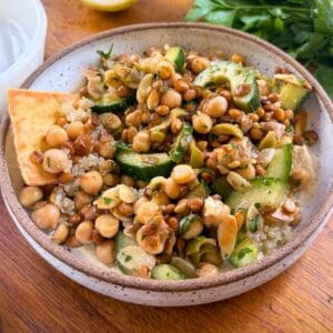 Brown-rimmed white bowl filled with lentils and chickpeas on a wood table with parsley and half of a lemon in the background.