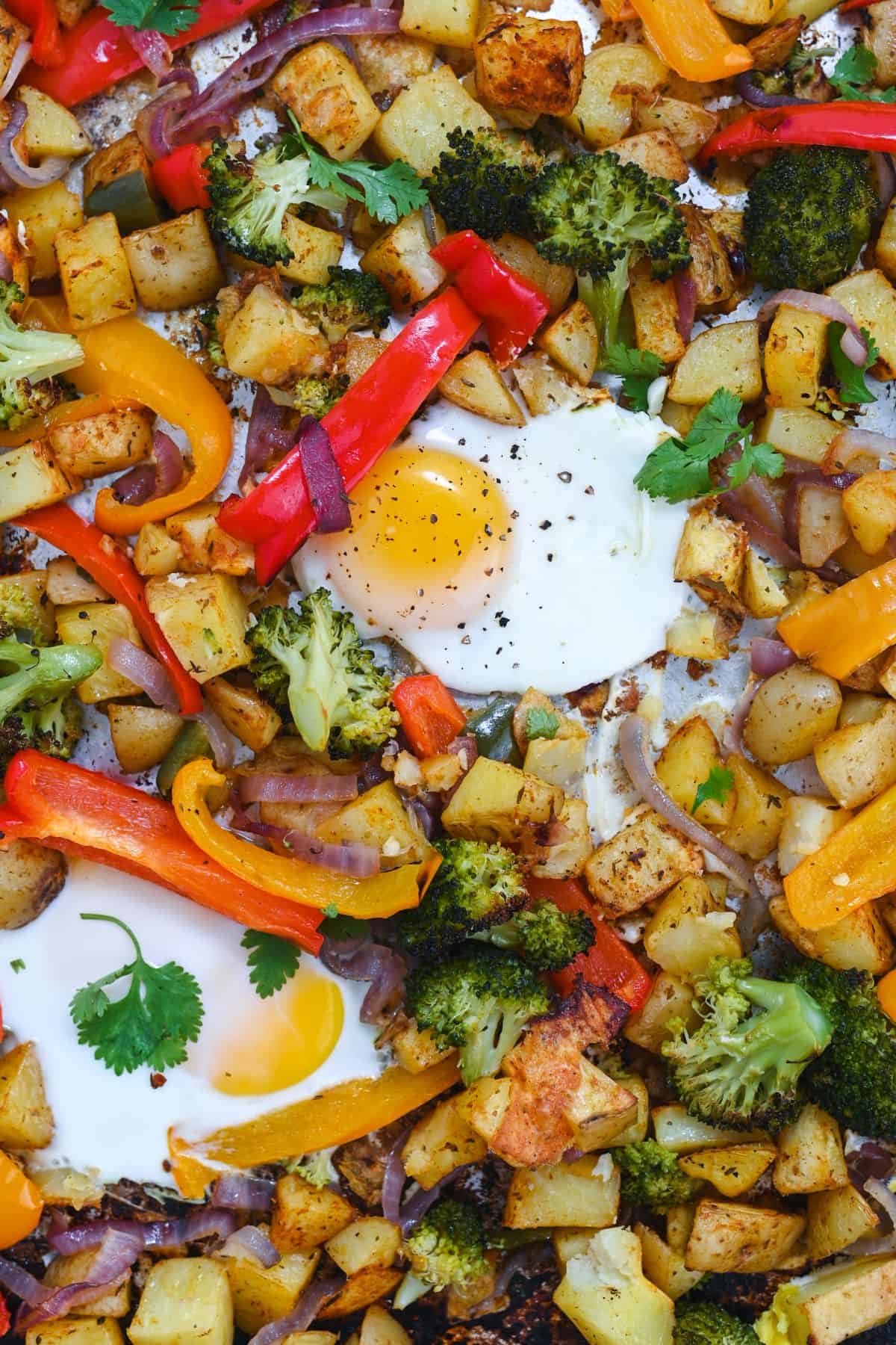 Close up of two fried eggs nestled among peppers, broccoli, onions, and potatoes.