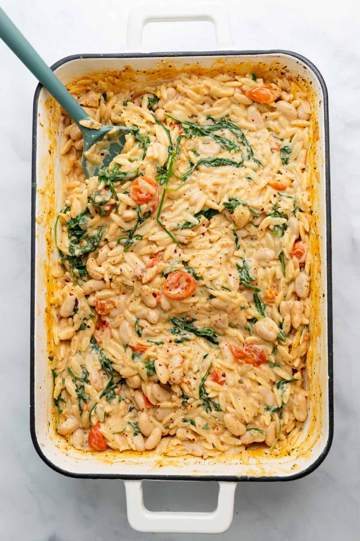 Overhead view of rectangular enamel baking dish filled with creamy orzo and a green serving spatula.