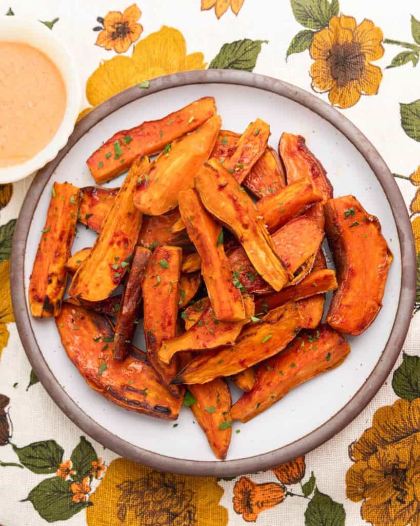 Sweet potato wedges piled on a brown-rimmed white plate on a colorful flower tablecloth.
