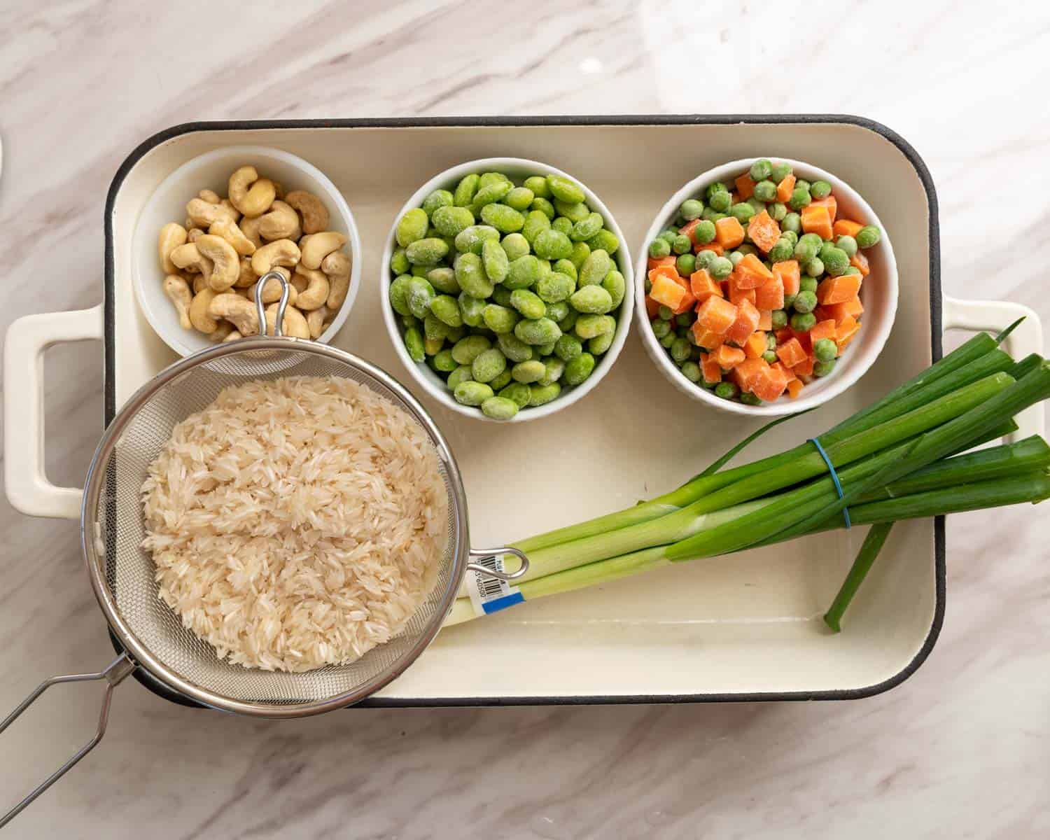 White rectangular enamel casserole dish with green onions and bowls of cashews, mukimame, frozen peas and carrots, and rinsed white rice.