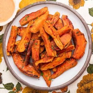 Sweet potato wedges piled on a brown-rimmed white plate on a colorful flower tablecloth.