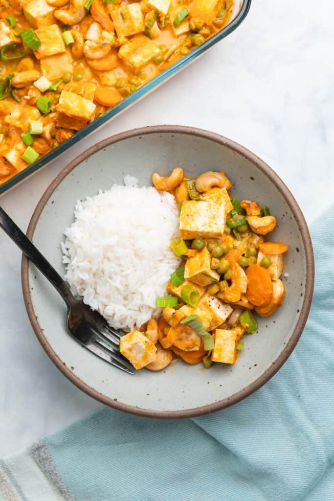 Gray bowl filled with rice, tofu curry, and a black fork with a glass baking dish of curry in the top left corner.
