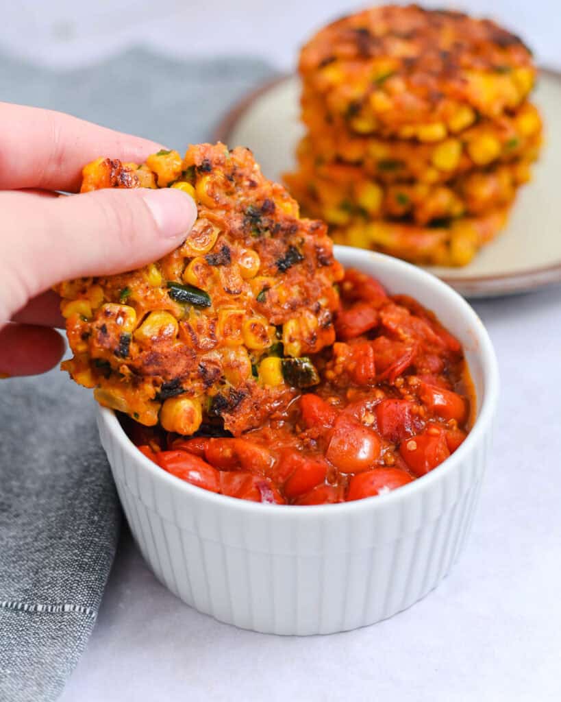Corn fritter being dipped into a ramekin of homemade cherry tomato sauce.