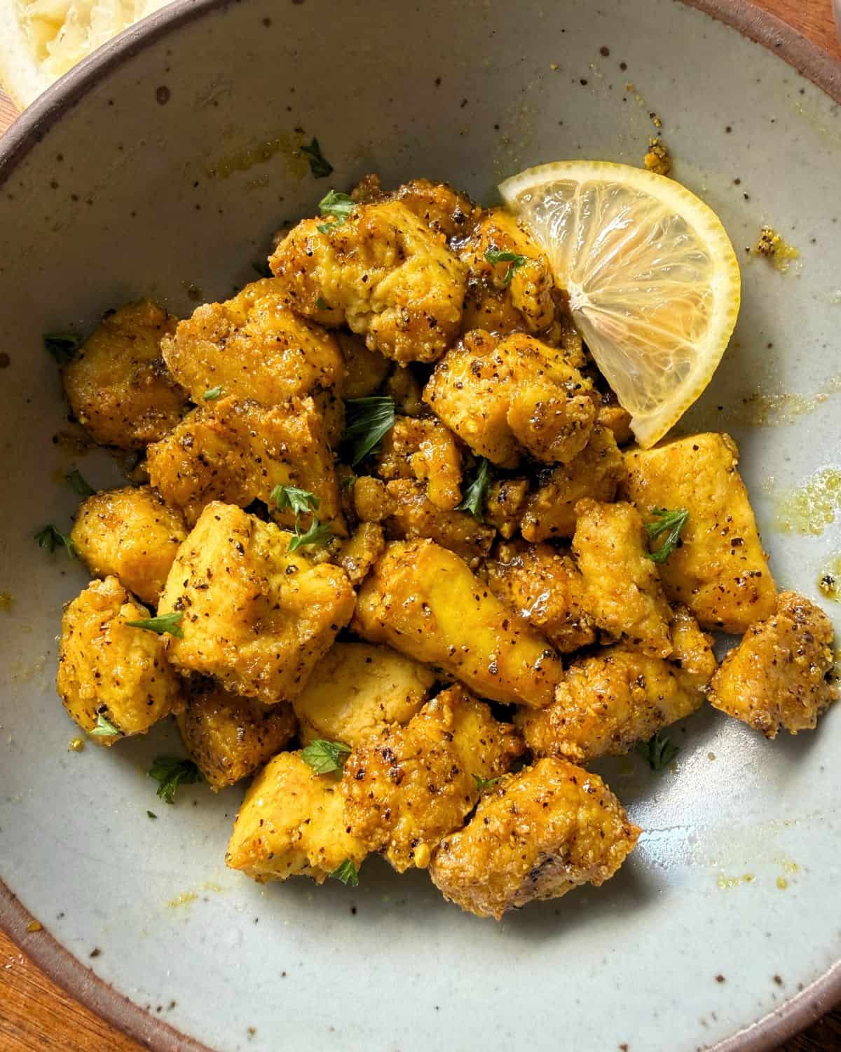 Gray bowl with a wedge of lemon and crispy nuggets of tofu in lemon pepper sauce.