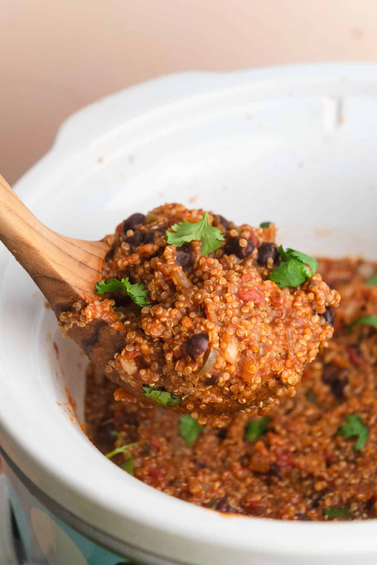 Wood ladle lifting a scoop of seasoned quinoa out of a white slow cooker pot.