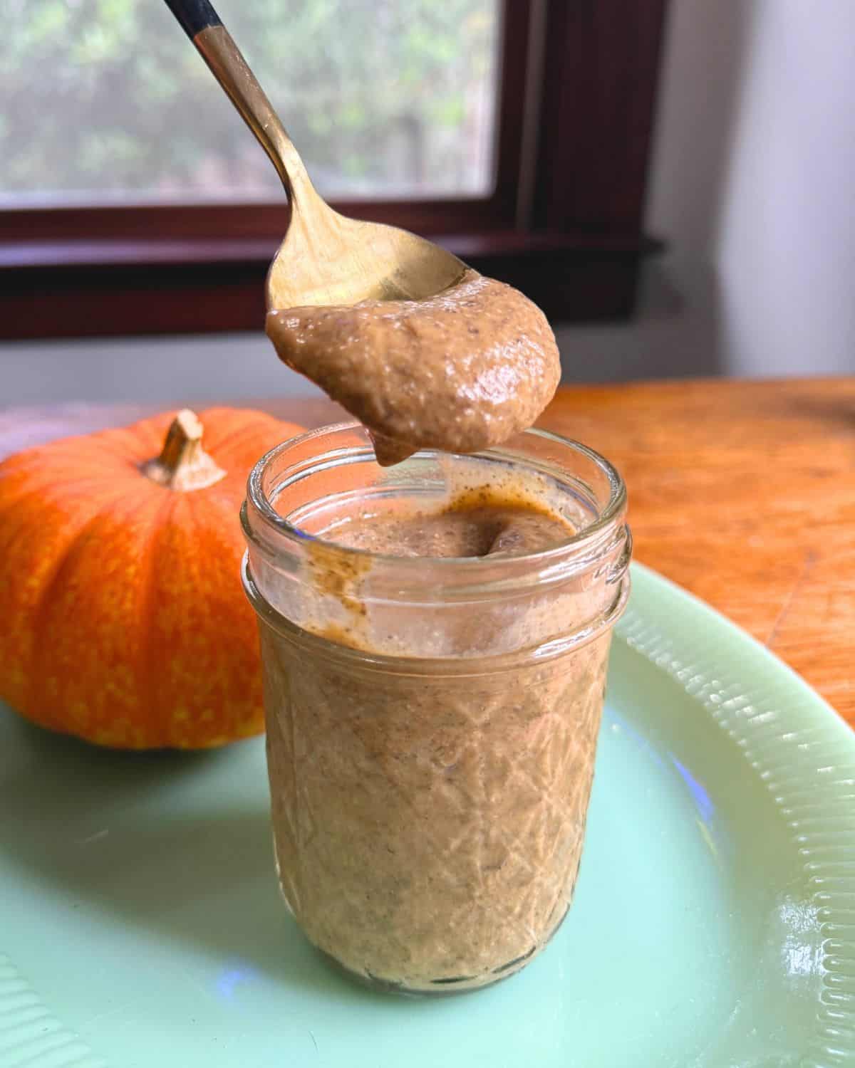 Small mason jar with chia pudding on a jade plate with an orange pumpkin in the background and a spoonful of chia pudding hovering above the jar.