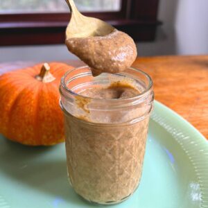 Small mason jar with chia pudding on a jade plate with an orange pumpkin in the background and a spoonful of chia pudding hovering above the jar.