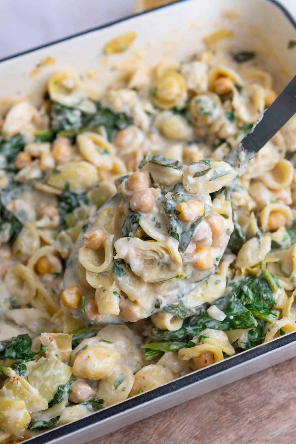 Metal serving spoon lifting a portion of pasta out of a white enamel baking dish.