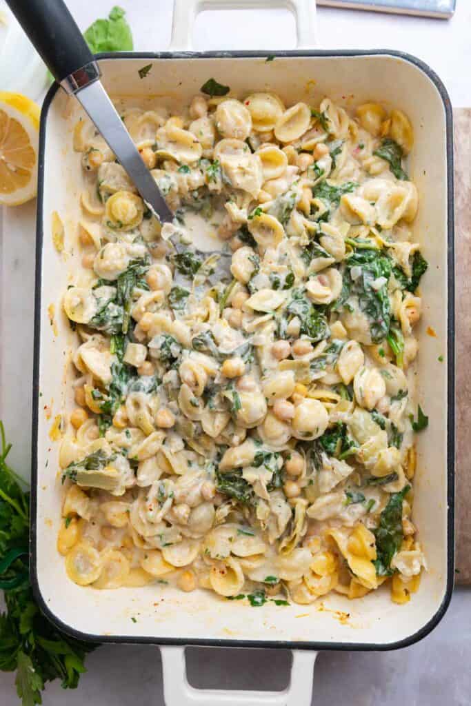 Overhead view of spinach artichoke pasta in a white enamel baking dish with a serving spoon.