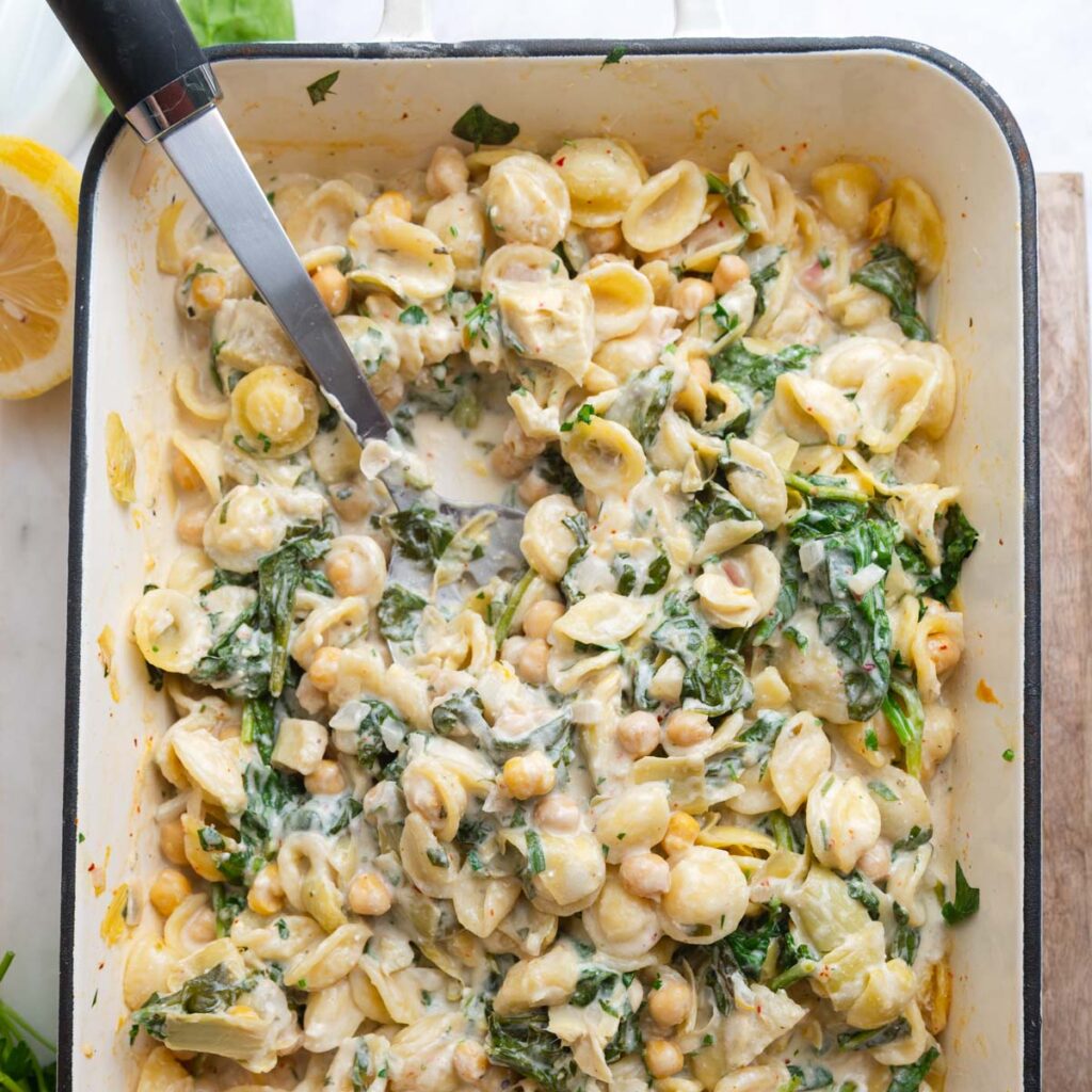 Overhead view of spinach artichoke pasta in a white enamel baking dish with a serving spoon.